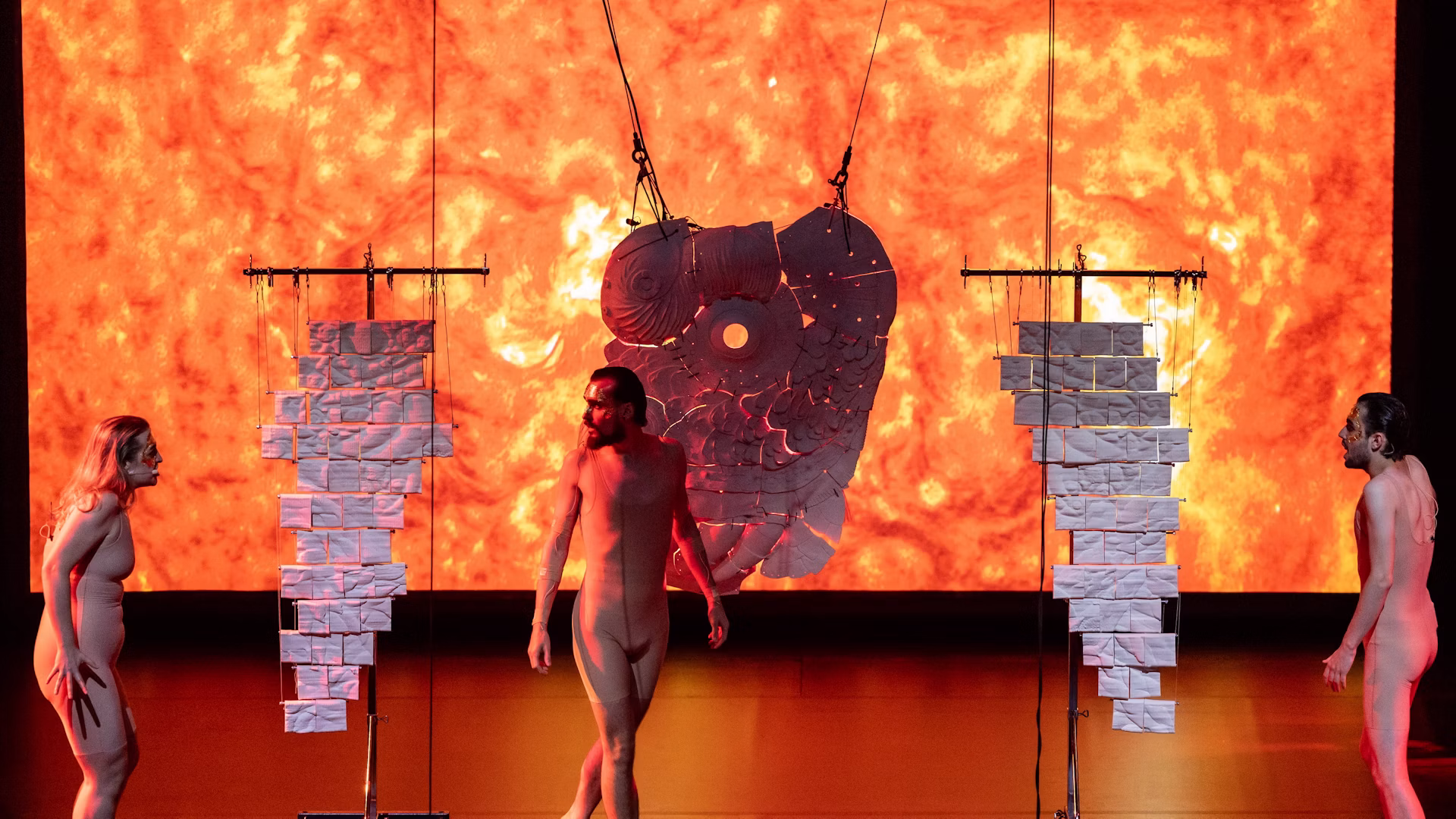 Three performers stand before the central orange solar projection in Graz.